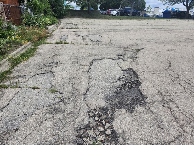 An asphalt parking lot with large cracks, pothole and weeds growing, with vehicles in another lot off in the distance.