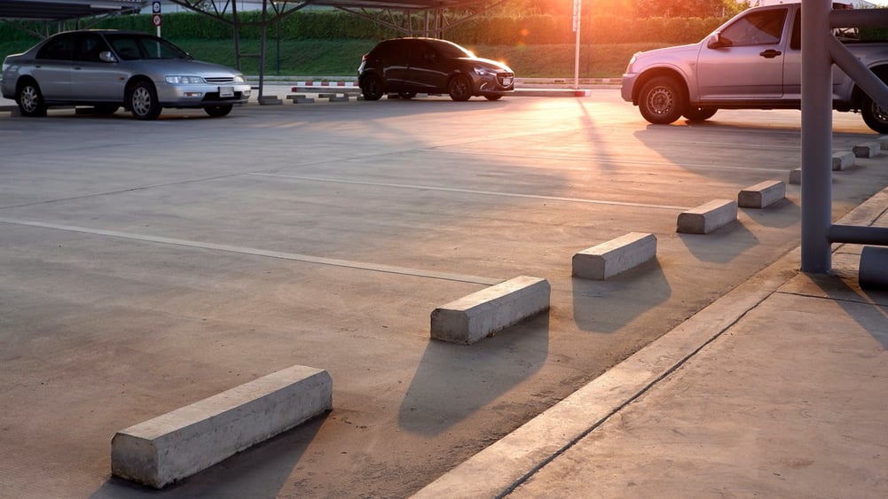 A smooth, light-colored concrete parking lot at sunset. The foreground shows several concrete parking blocks and painted space lines, while cars are parked under a covered structure in the background.