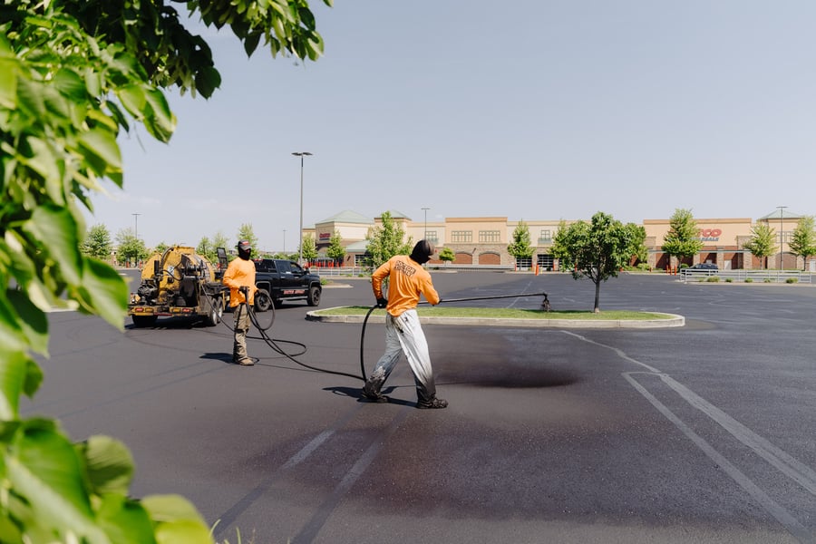 Two Doctor Asphalt crew members in bright orange shirts using professional spray equipment to apply a precise, even layer of sealcoat across a large commercial parking lot on a clear day.