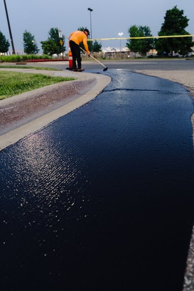 A close-up view of freshly applied, smooth black sealcoat neatly edging a concrete curb, with a Doctor Asphalt professional carefully applying sealant in the background to ensure even coverage.