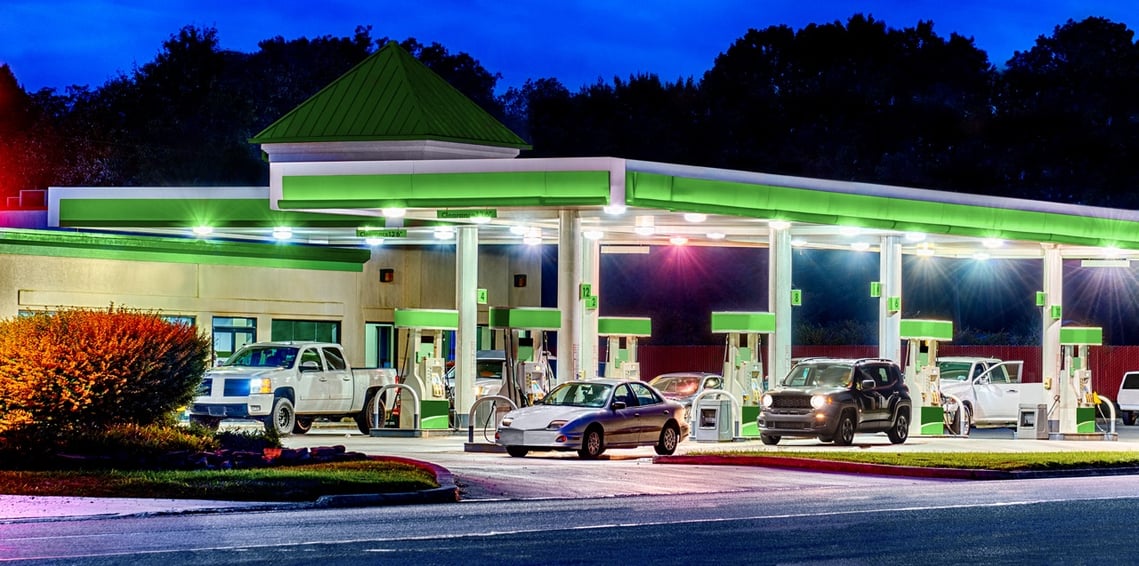 A lit up gas station at night with green accents and pumps with several cars and trucks parked in paved parking lot with frontage road close by.