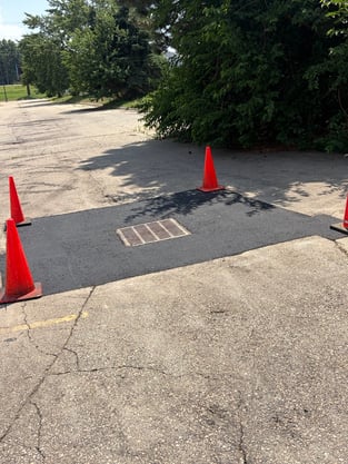 A seamless, square patch of new asphalt surrounding a metal storm drain grate in a parking lot, marked by four orange traffic cones to demonstrate an infrared asphalt repair.