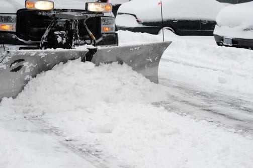 white snow plow truck pushing several inches of snow in a parking lot with cars parked in the background
