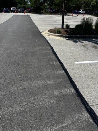 A long strip of freshly laid, dark asphalt in a parking lot, showing a distinct, sealed seam against the older, faded pavement to illustrate a traditional cut-and-patch repair.
