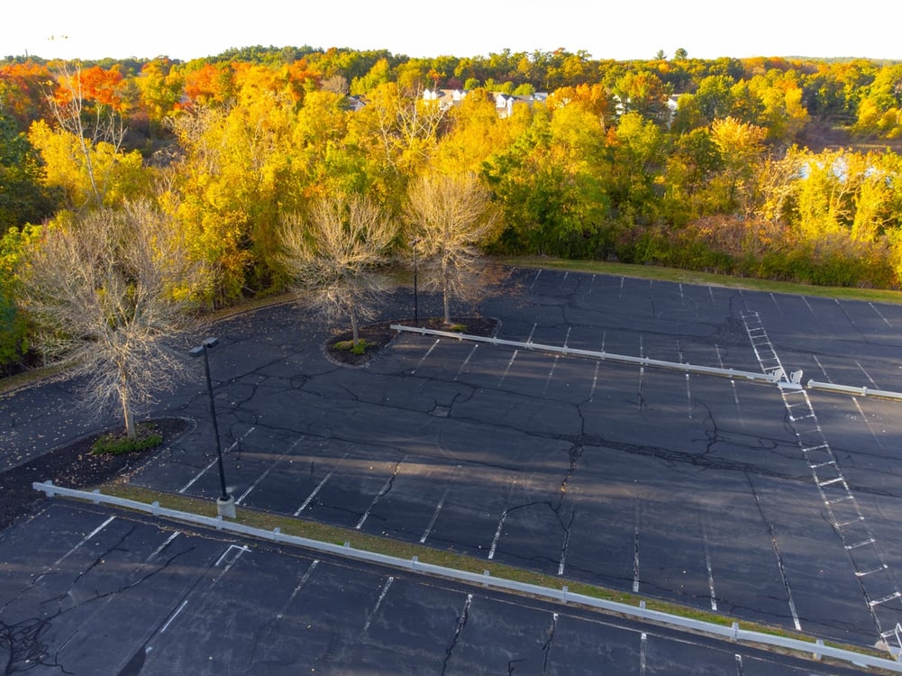 aerial view of a commercial parking lot with cracks and fade line striping among lots of colorful trees during the fall season