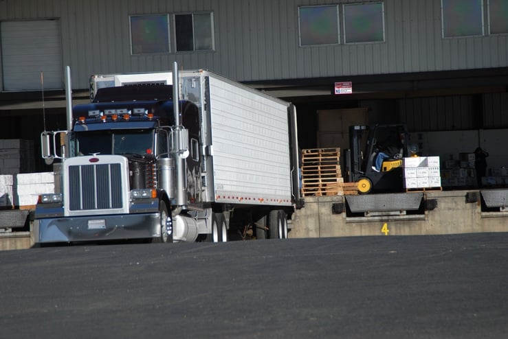 a large, heavy semi truck parked on black asphalt pavement close to load dock area with pallets and forklift