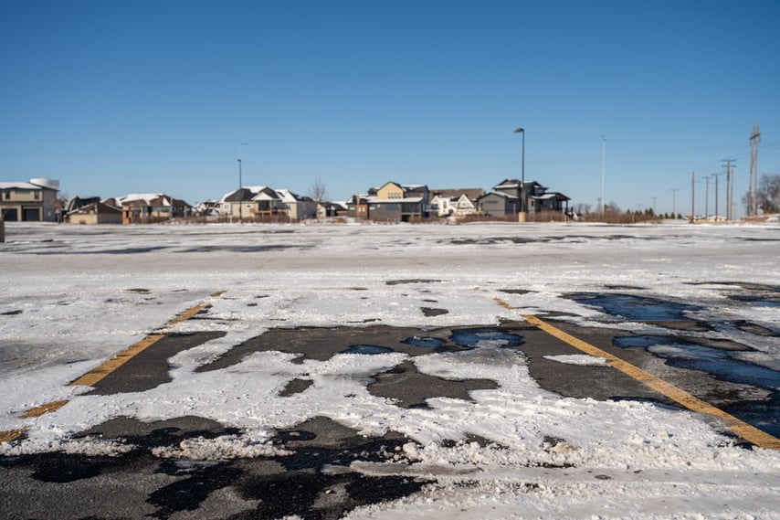 A snow covered parking lot with the lines covered in patchy icy and snow in several locations with houses in the distance background