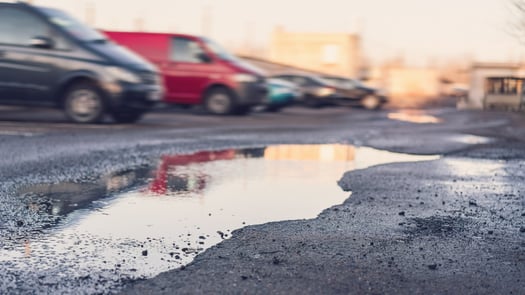 A large puddle reflecting the sunset on a worn asphalt parking lot surface, with blurred vehicles parked in the background.