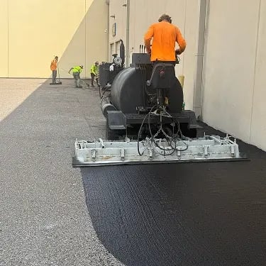 Doctor Asphalt crew applying microsurfacing with spreader box along building perimeter
