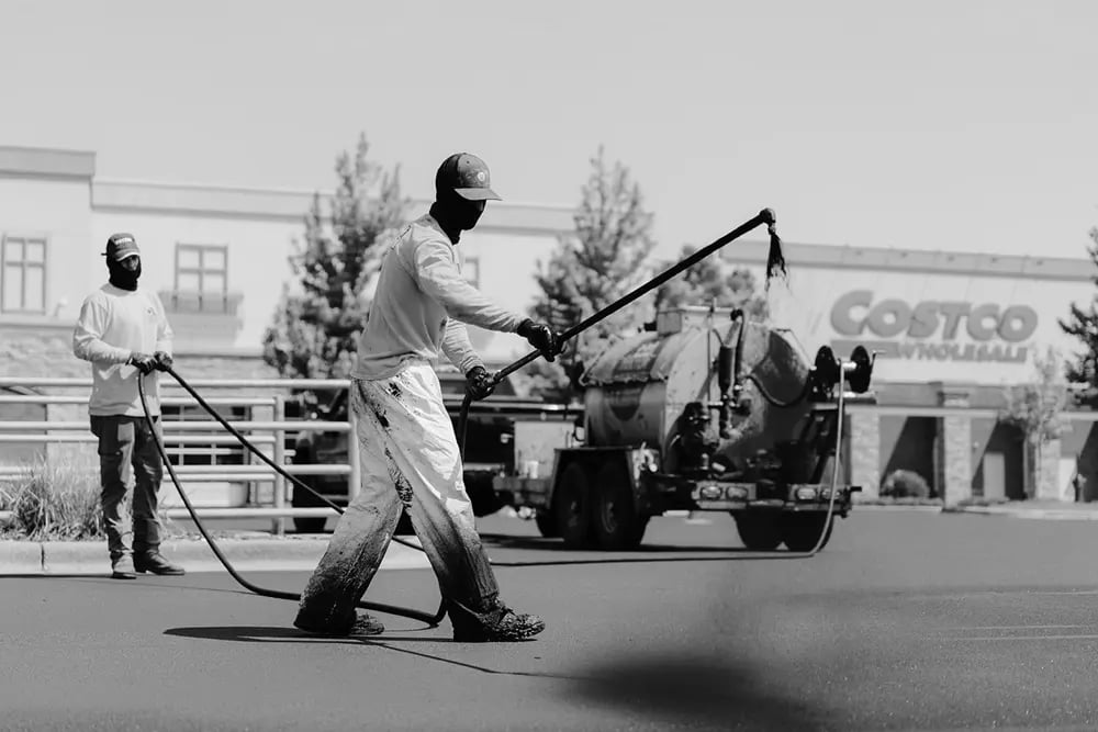 Doctor Asphalt crew applying sealcoat spray to parking lot pavement with tank sprayer