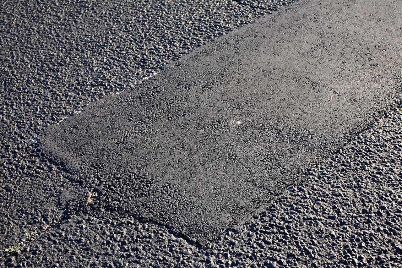 A close-up view of a rectangular patch of smooth, newly laid asphalt contrasting with the rougher texture of the surrounding older pavement in a parking lot.