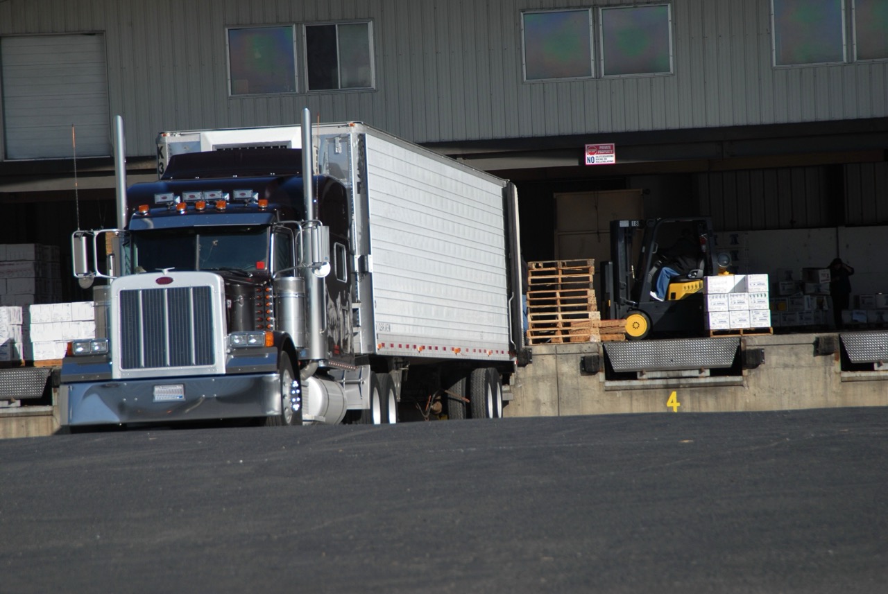 a large, heavy semi truck parked on black asphalt pavement close to load dock area with pallets and forklift