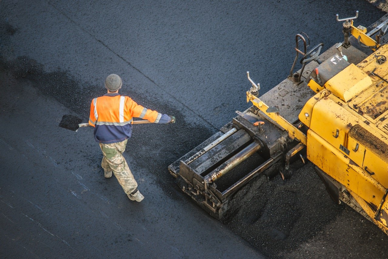 asphalt company employee laying down fresh asphalt pavement in commercial parking lot with heavy equipment